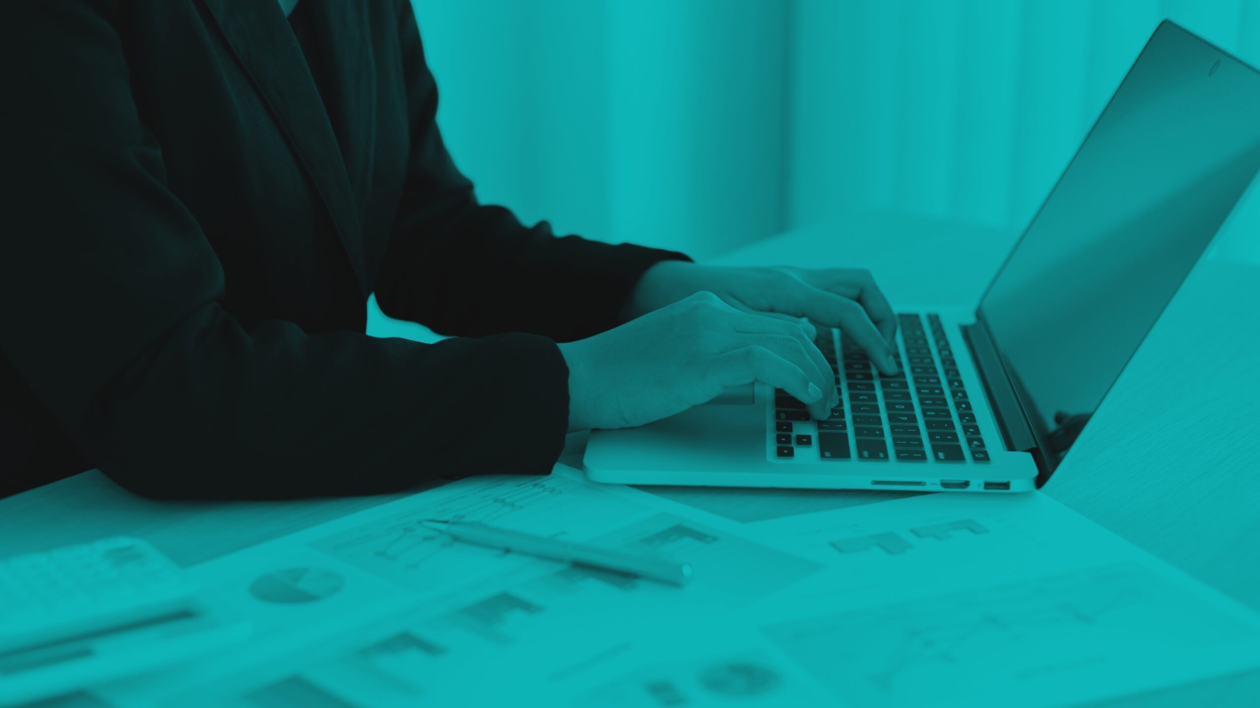 Person typing on a laptop with documents and charts spread out on the desk.
