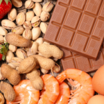 A variety of foods arranged on a wooden surface, including fresh strawberries, pistachios in their shells, whole peanuts in their shells, cashew nuts, peeled shrimp, two brown eggs, and two bars of milk chocolate.