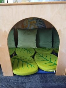 A cozy reading nook made of light-colored wood with an arched opening. Inside, there are three green square pillows against the back wall and several round green cushions with leaf patterns arranged on the floor.