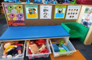 A classroom reading and play area featuring a blue cushioned bench with a green plastic chair attached on the right side. Under the bench are three clear bins filled with toys, including dolls, plush animals, and sensory items. Above the bench, a corkboard displays colorful posters, including one with cartoon pigeons, a library sign with the number 2, and charts about feelings and classroom rules.