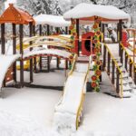 Snow-covered playground with slides, bridges, and climbing structures in a park.