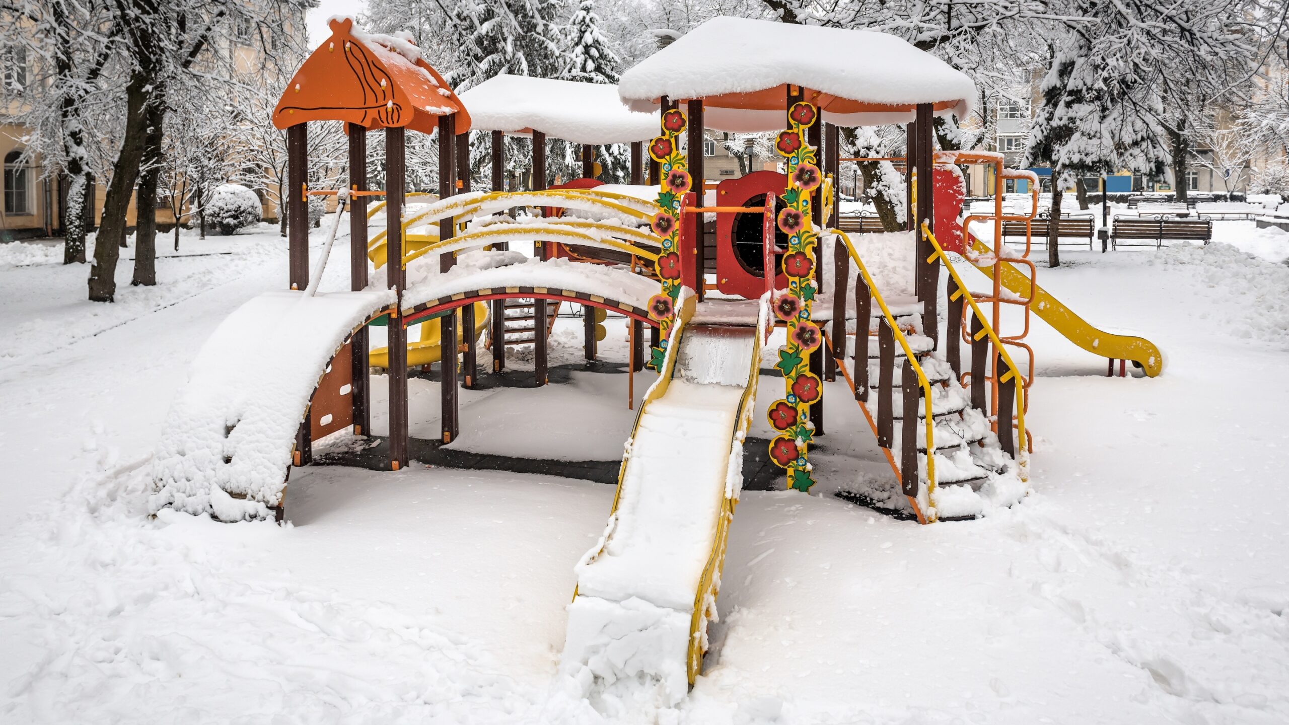 Snow-covered playground with slides, bridges, and climbing structures in a park.