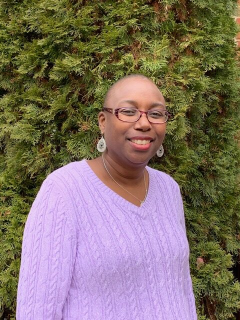 Person wearing a light purple cable-knit sweater standing in front of green shrubs.