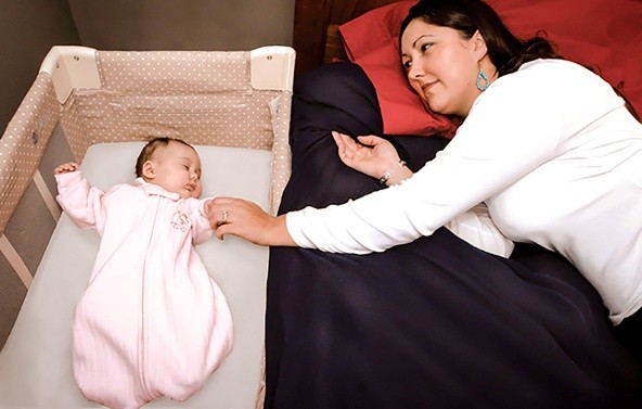 Adult lying in bed next to a bedside bassinet with an infant in a pink sleep sack.