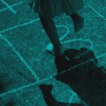 Feet stepping on a chalk-drawn hopscotch grid on pavement, with a shadow cast on the ground.