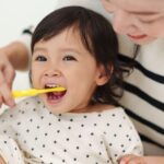 Adult helping a young child brush their teeth while sitting together.