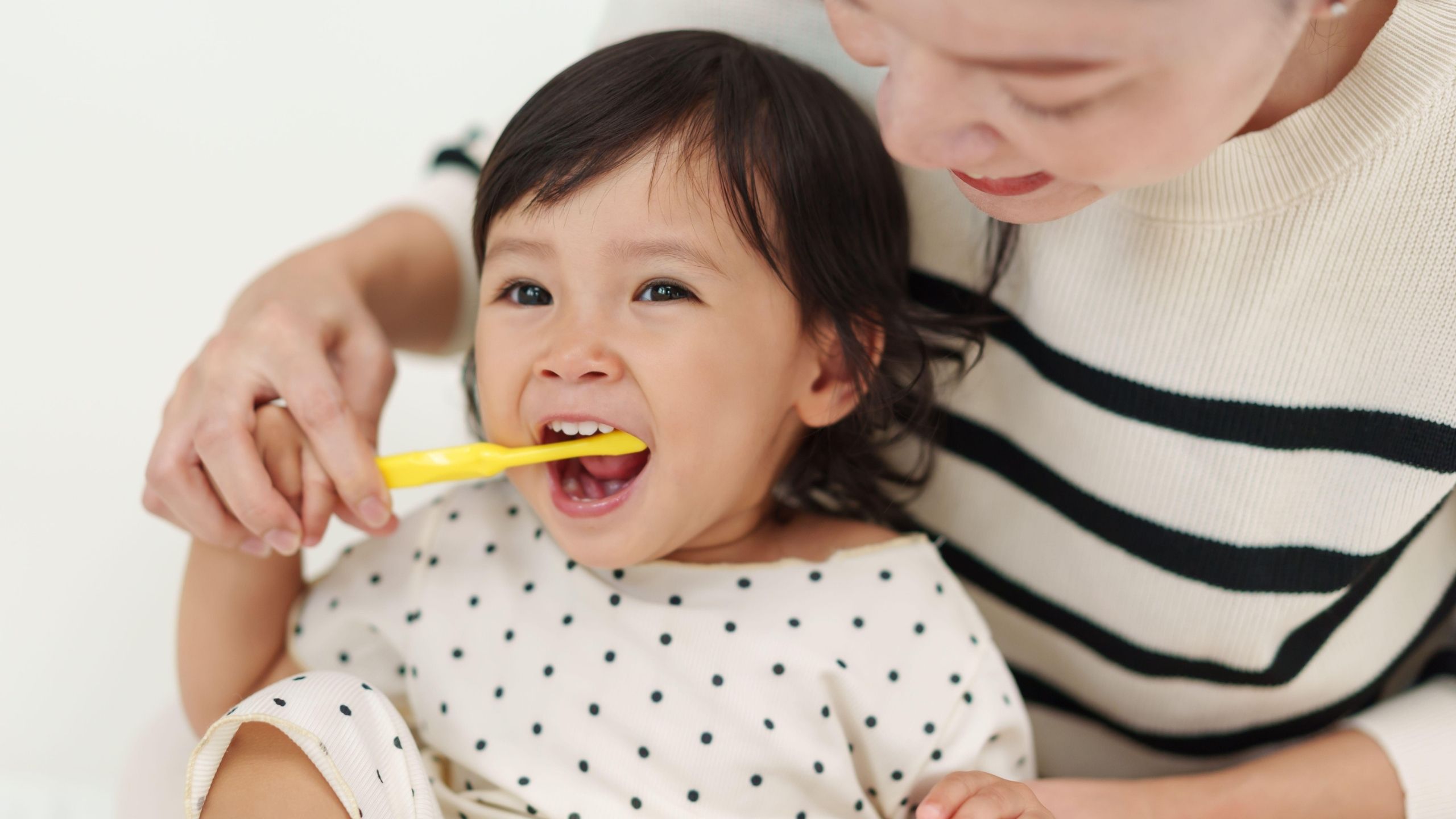 Adult helping a young child brush their teeth while sitting together.