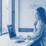 Person sitting at a desk near a large window, working on a laptop with a smartphone placed beside it. Shelves with books and documents are visible in the background.