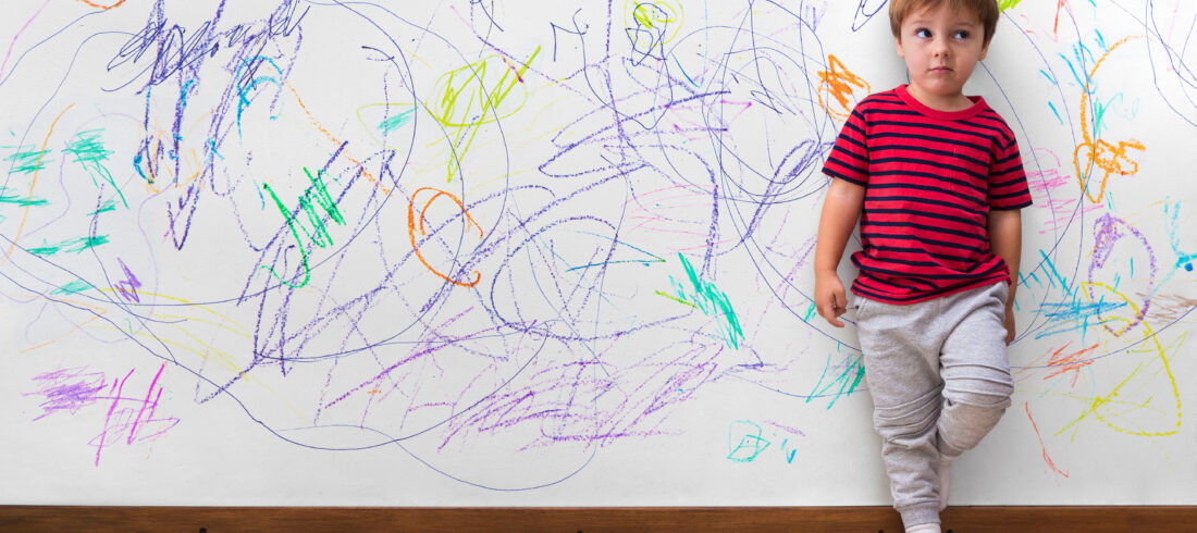 Young child standing against a wall covered in colorful crayon scribbles, with crayons scattered on the floor.