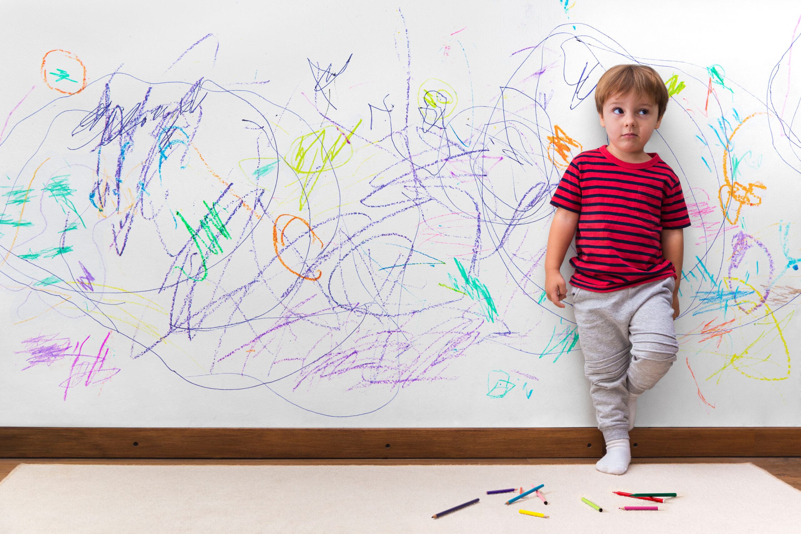 Young child standing against a wall covered in colorful crayon scribbles, with crayons scattered on the floor.
