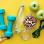 Assorted fitness and healthy lifestyle items on a yellow background, including bananas, green apples, dumbbells, a water bottle, nuts, a jump rope, and a bowl of oats.