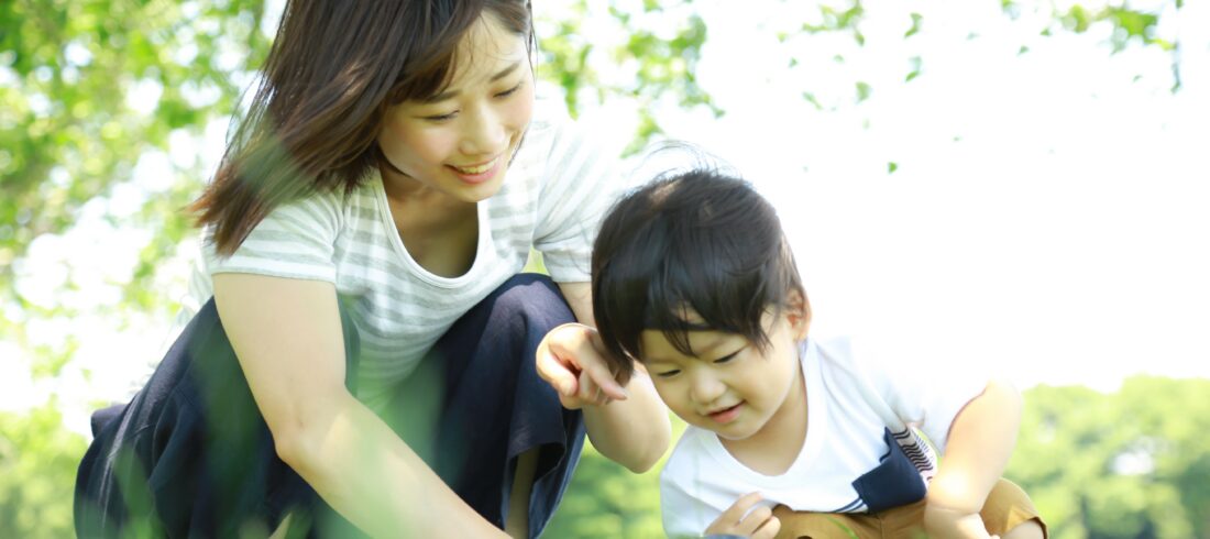 An adult and a child crouching in the grass outdoors, using a magnifying glass to look closely at something on the ground.