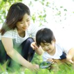 An adult and a child crouching in the grass outdoors, using a magnifying glass to look closely at something on the ground.