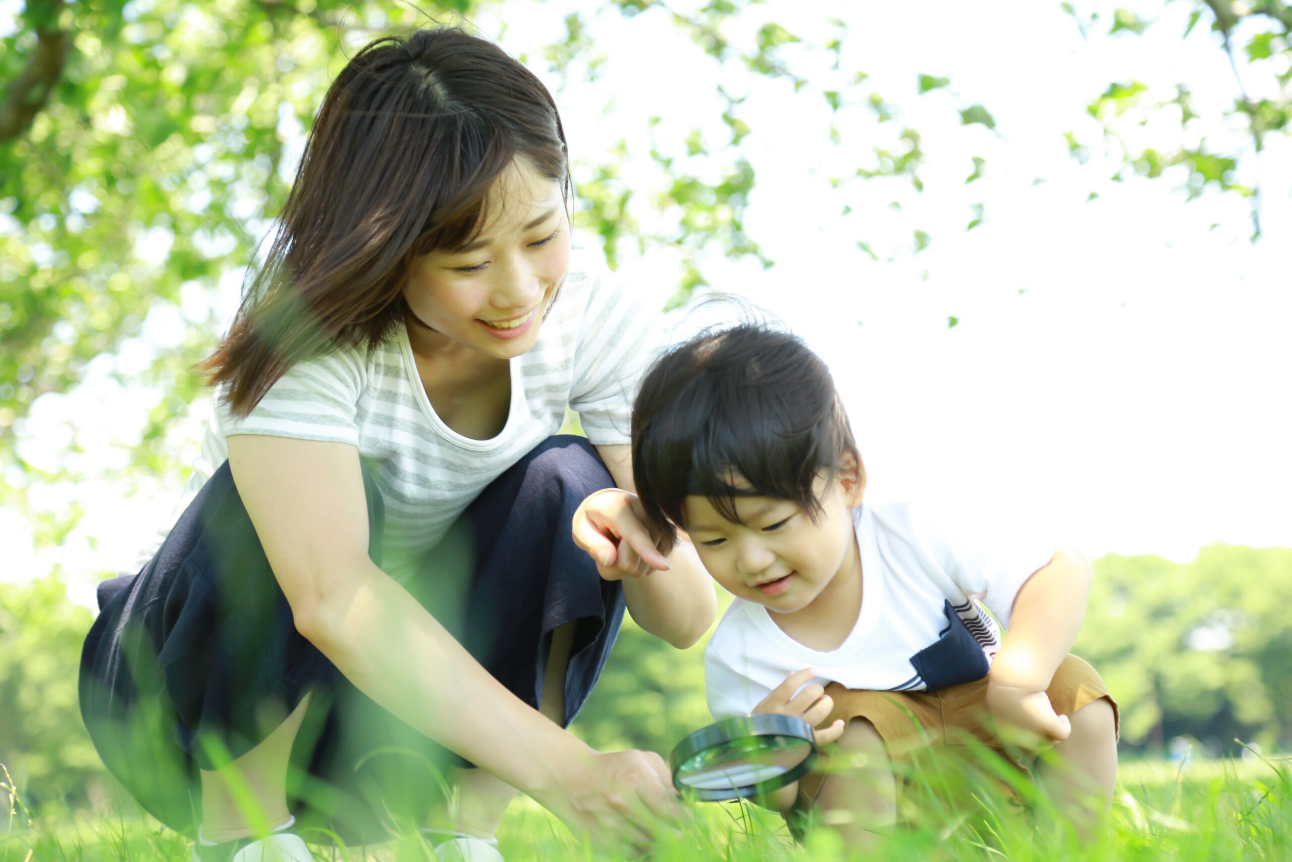 An adult and a child crouching in the grass outdoors, using a magnifying glass to look closely at something on the ground.
