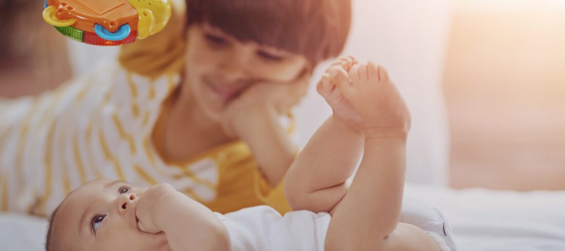 An older child holding a colorful toy engages with a baby lying on a bed in soft, warm lighting.