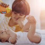An older child holding a colorful toy engages with a baby lying on a bed in soft, warm lighting.