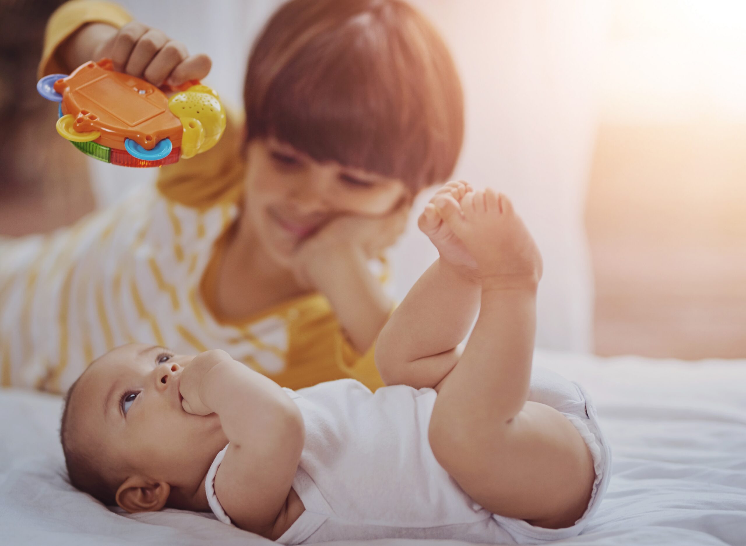 An older child holding a colorful toy engages with a baby lying on a bed in soft, warm lighting.