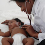 Healthcare professional using a stethoscope to examine an infant lying on a soft surface.