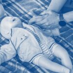 Hands practicing infant CPR on a training mannequin laid on a blanket.