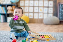 Young child sitting on the floor, holding a pink toy in the mouth, with wooden toys scattered nearby in a playroom.