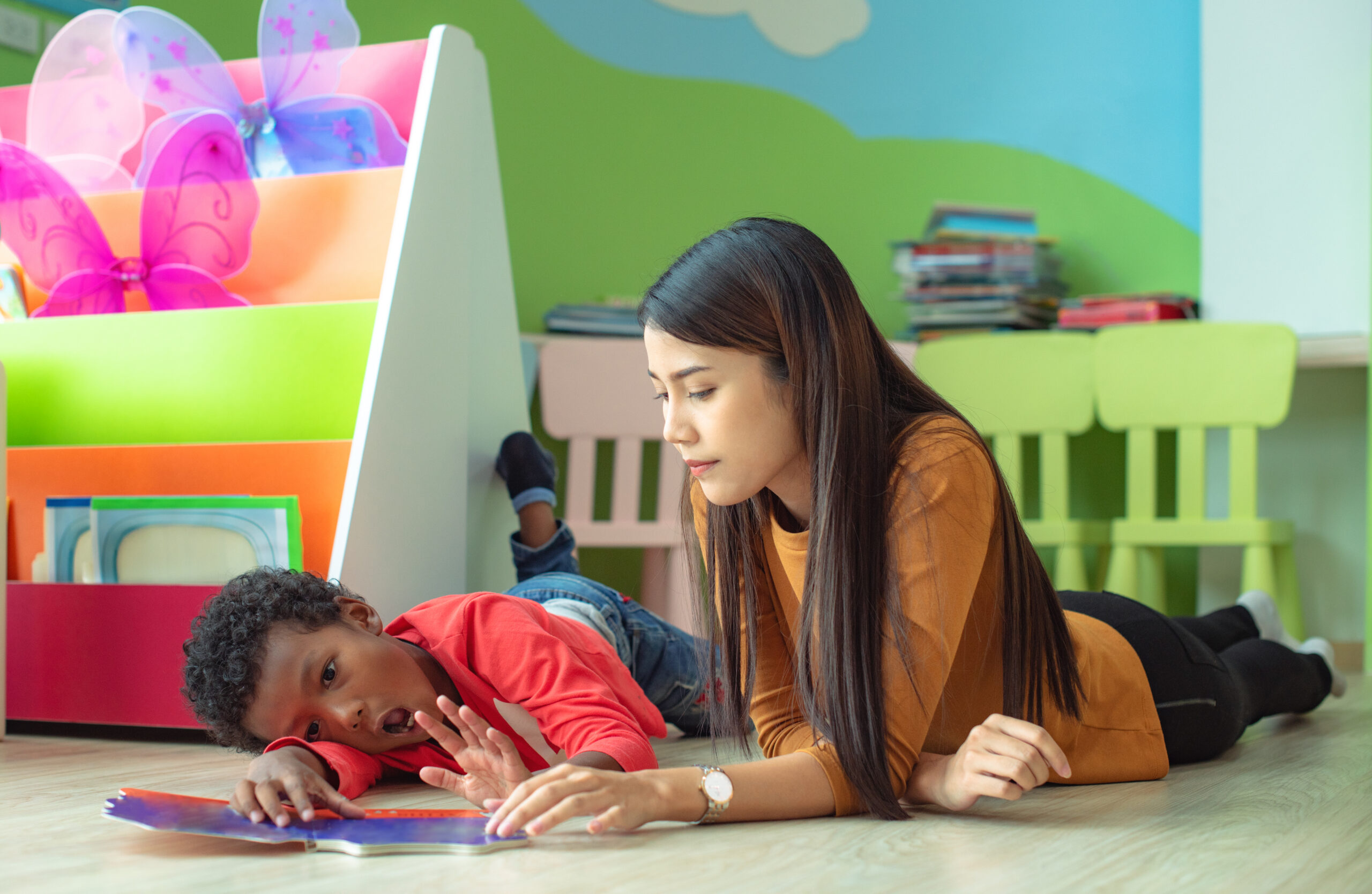 Adult and child lying on the floor reading a book together in a colorful early‑learning classroom.