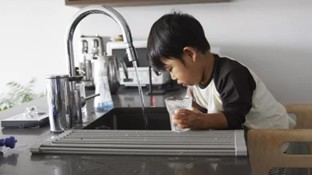 Small child standing at a kitchen sink, drinking water from a clear cup while leaning on the counter.