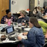 Participants sit at round tables in a conference room, collaborating in small groups with laptops, papers, and notes during a workshop or training session.