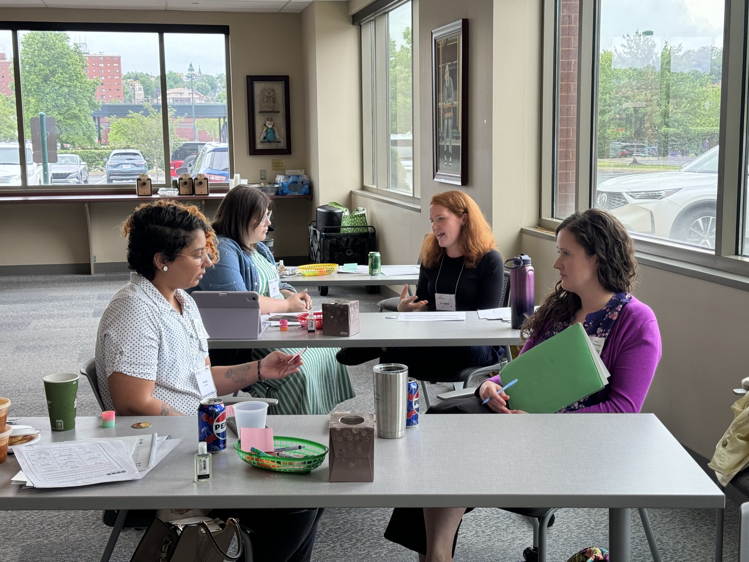 Small group workshop setting with participants seated at a table, discussing materials and taking notes with folders, tablets, and drinks near large windows.
