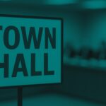 Town hall sign in the foreground with a blurred meeting table and seated council members in the background.