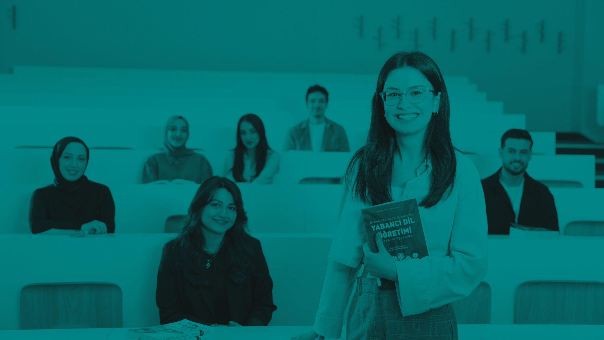 Teal‑filtered image of a student standing in a tiered classroom holding a book, with other students seated in the background.