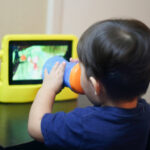 Young child sitting at a table drinking from a cup while watching a video on a tablet in a bright yellow protective case.