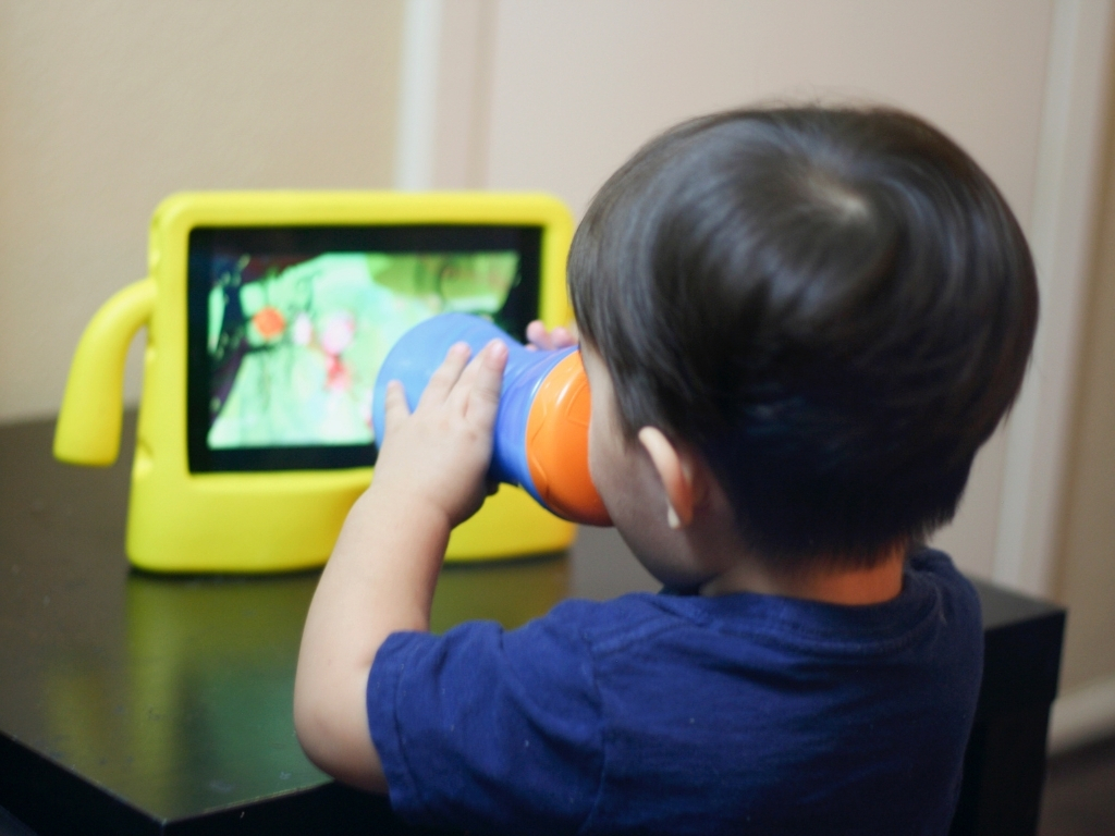 Young child sitting at a table drinking from a cup while watching a video on a tablet in a bright yellow protective case.