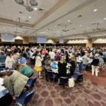 Large conference room filled with attendees standing around tables during a group activity, with presentation screens visible at the front.