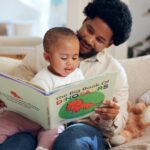 Adult and young child sitting on a couch reading a large picture book together at home.